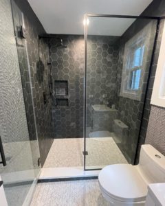 A modern bathroom showcasing a walk-in shower with dark gray hexagonal tiles, complemented by lighter patterned tiles on the floor. The shower is enclosed with a sleek, clear glass panel and features a built-in niche and a contemporary shower head. The rest of the space includes a white toilet, and the design is unified by a muted gray color scheme.