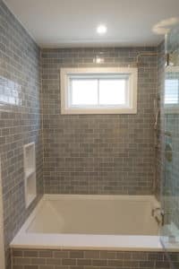 A sophisticated bathroom featuring a combination of bathtub and walk-in shower. The walls are adorned with grey subway tiles and complemented by a glass shower partition. The bathtub is recessed with a wide ledge for added convenience. A window provides natural light, and the shower boasts modern fixtures, including a rainfall shower head.