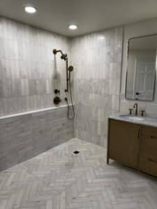 This image features a spacious and elegantly designed bathroom with white tiles covering the walls and floor in a herringbone pattern. It includes a luxurious shower setup with bronze fixtures, including a handheld showerhead, and well-placed lighting that highlights the sleek and clean design.