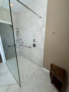 This image features a modern shower with light gray walls, white pebble floor tiles, and chrome fixtures, enclosed by a glass panel. A wooden shower seat adds a warm touch.